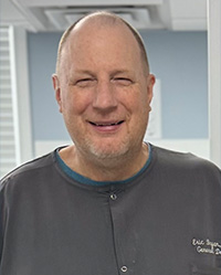 The image shows a man with a bald head, wearing glasses and a blue shirt, standing in front of what appears to be a medical or dental office setting.