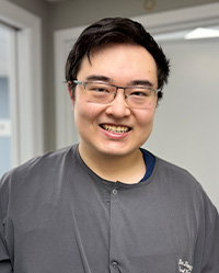 Man with glasses and a beard, smiling at the camera, standing in an indoor setting that appears to be a professional office or waiting area.