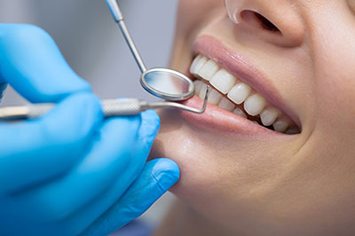 The image depicts a person receiving dental care, with a dental professional using a drill on their teeth while the patient is wearing a blue surgical gown and a hairnet.