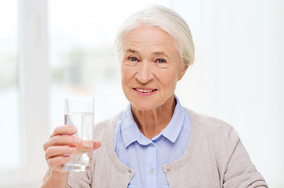 The image is a photograph of an elderly woman holding a glass of water, smiling, and looking directly at the camera.