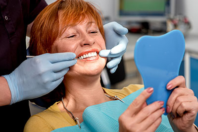 The image shows a woman sitting in a dental chair, receiving dental care. She is holding a blue dental impression tray and smiling at the camera.