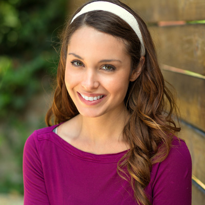 A smiling woman with long brown hair, wearing a purple top and headband, poses against a wooden fence.