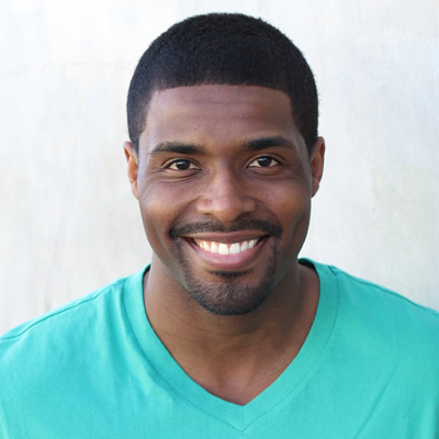 An elderly couple is smiling and embracing each other, with the man on the left wearing a patterned shirt and the woman on the right wearing a blue scarf. They are seated outdoors, possibly in a park or garden setting.