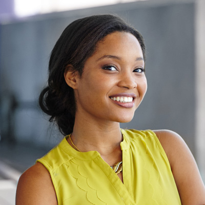 A smiling woman with dark hair, wearing a yellow top and standing in front of a wall.