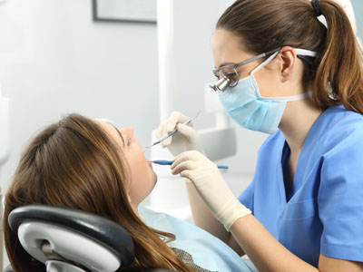 A dental hygienist is performing a teeth cleaning procedure on a woman, with the dental hygienist wearing protective gear and using dental tools.