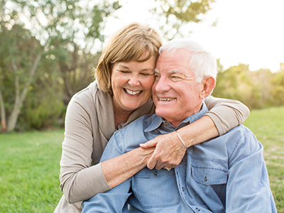 An elderly couple embracing outdoors, with the man seated and the woman standing behind him.