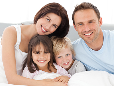 The image shows a family of four, including two adults and two children, lying together in bed with smiles on their faces.