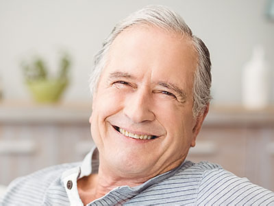 The image shows an older man with gray hair, smiling at the camera while sitting in a relaxed position.