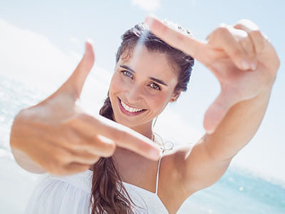 A smiling woman with long hair, holding up a peace sign in front of her face against a bright blue sky background.